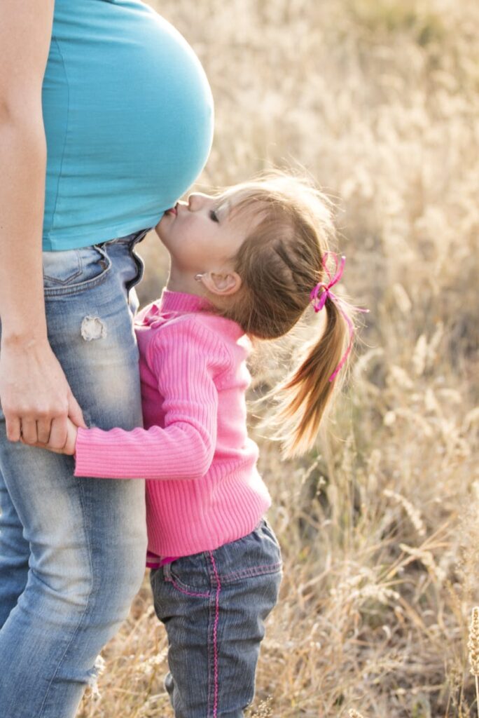 A young girl lovingly kisses her pregnant mother's belly outdoors, symbolizing family affection.